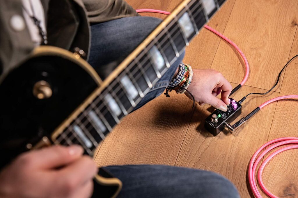 A guitarist adjusting the IK Multimedia ToneX One pedal while playing a black electric guitar, connected with pink cables.